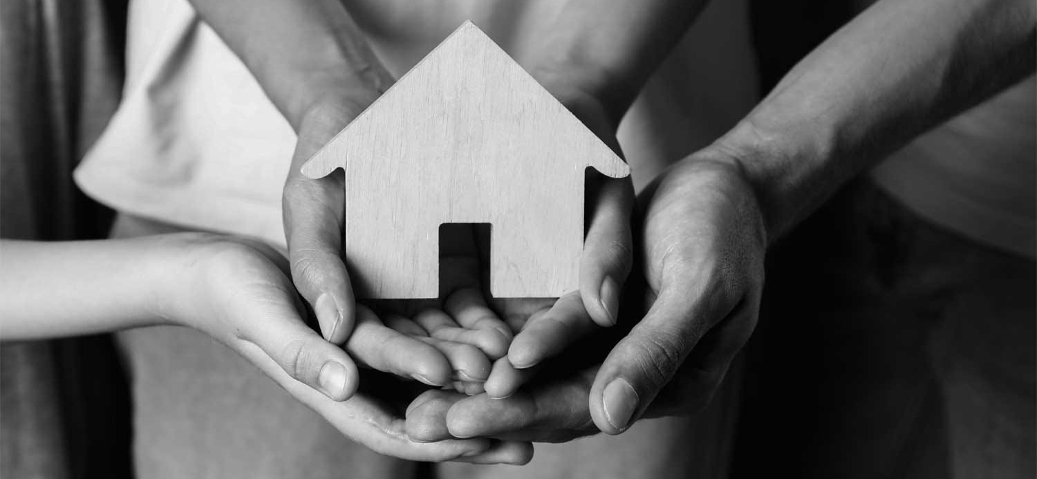 Image of a wooden block shaped like a house being held in the hands of three people.