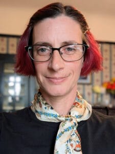 Image of Bekki in their home office. Bekki has short red hair and glasses. They are wearing a black shirt and a multicolored scarf.